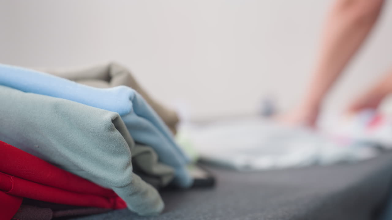 Colorful fabrics stacked neatly on table with blurred image of tailor arranging materials in background, showcasing creative preparation process in bright workspace filled with vibrant cloth pieces