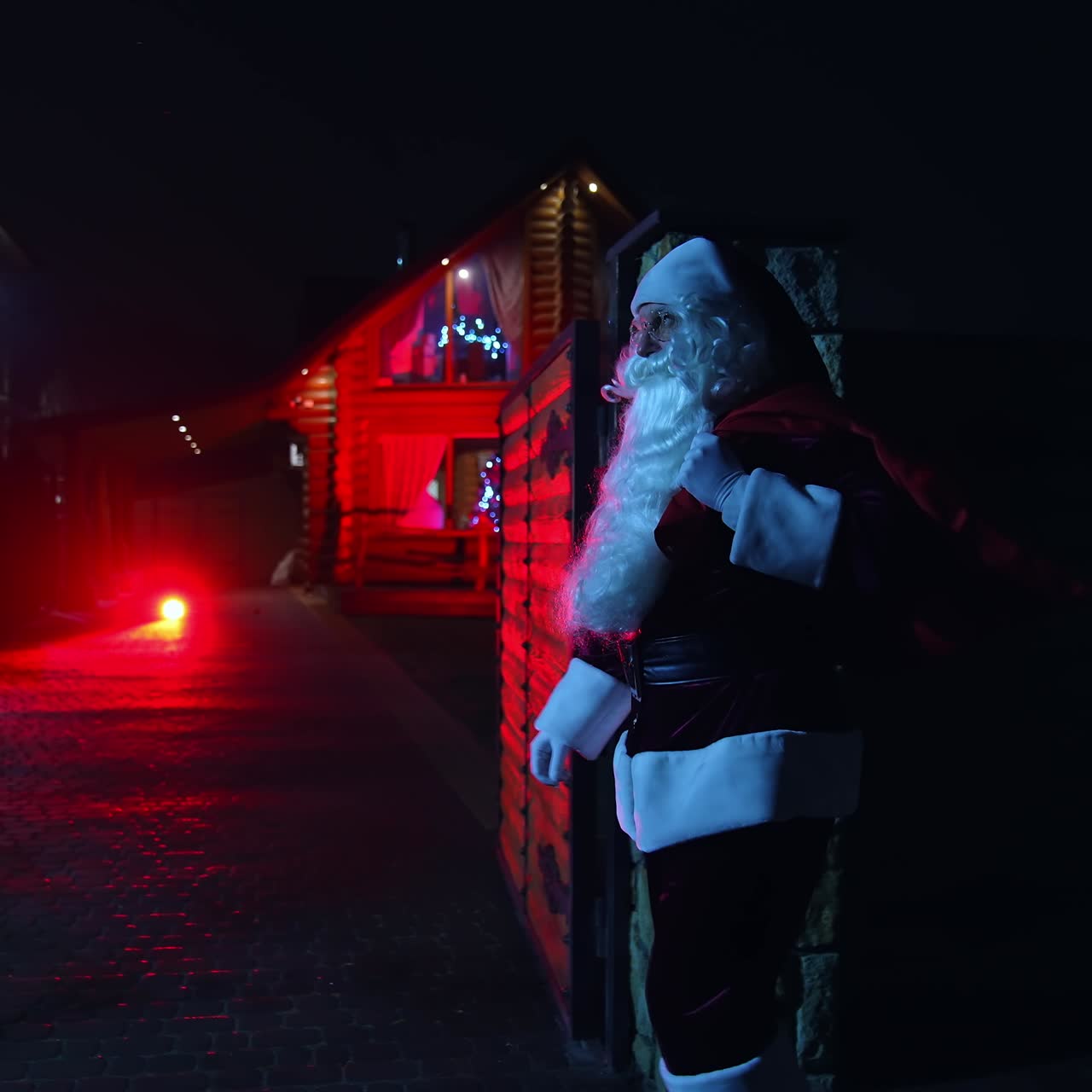 Santa Claus with red bag at night. Santa walks into front yard of house decorated with lights and garlands. Santa carrying gifts for children