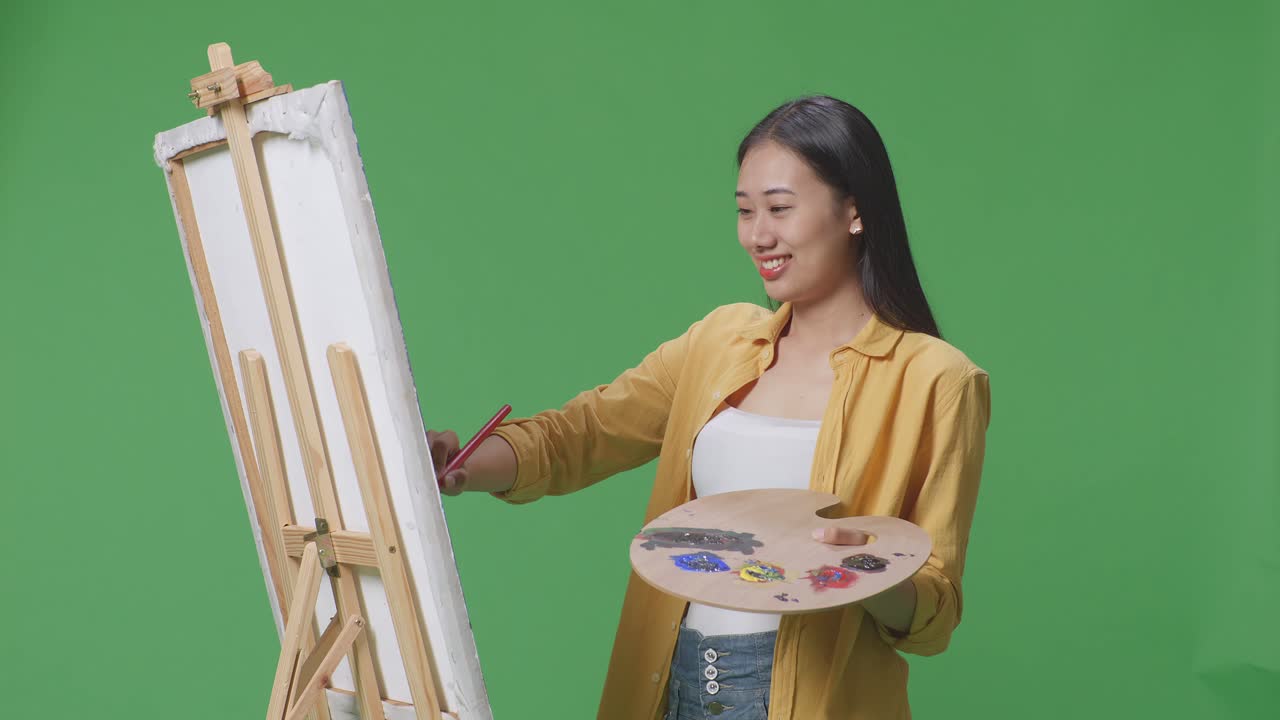 아시아 여성 예술가가 미소 짓고 카메라에 평화의 몸짓을 보여주면서 녹색 스크린 스튜디오에서 오일 페인트와 으로 캔버스에 그림을 그리고 있습니다.