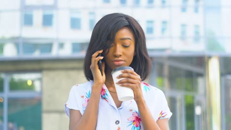 una joven afroamericana adulta está hablando por teléfono y bebiendo café afuera cerca de un edificio de oficinas moderno.