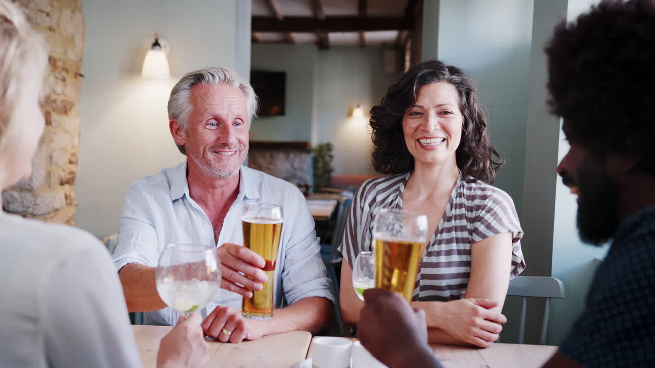 Senior white man and Hispanic woman sitting at a table in a pub making a toast with colleagues, close up, over shoulder view