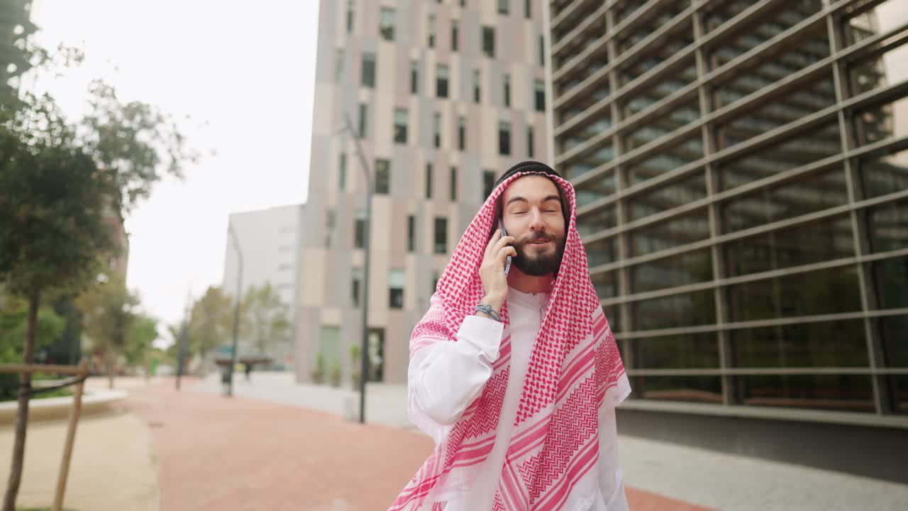 hombre árabe sonriente disfrutando de una conversación telefónica al aire libre, caminando por la calle de la ciudad en dubai