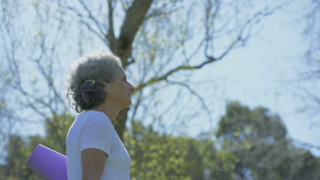 Side-view-of-woman-walking-in-park,-holding-yoga-mat-in-hand