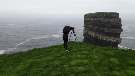 Female-photographer-with-a-tripod-and-digital-camera-takes-a-photo-of-a-huge-sea-stack-in-Ireland-at-Downpatrick-Head-on-the-edge-of-a-huge-cliff