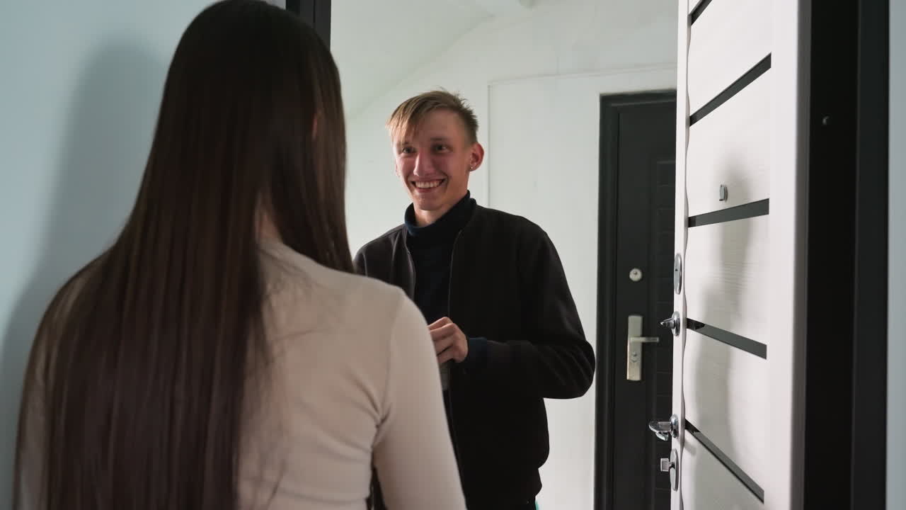 Una donna apre la porta al corriere, sorride e consegna un pacco sulla soglia dell'appartamento. Saluto rapido e consegna sicura ripresi da dietro, corridoio minimalista, design moderno della porta, luce naturale