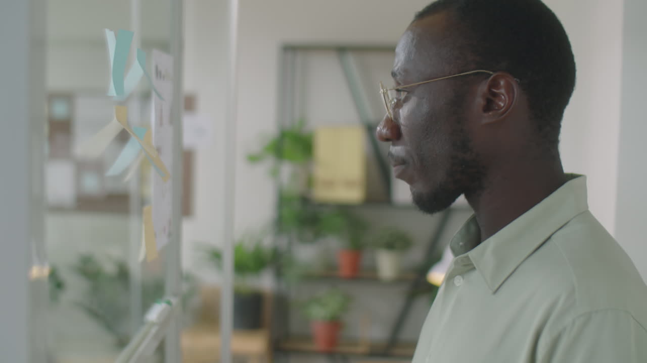 Black Businessman Writing on Sticky Notes on Glass Office Wall