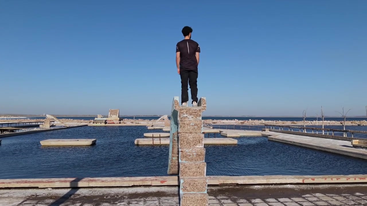 Pessoa sobe um escorregador aquático em ruínas para uma estrutura de piscina natural em Villa Epecuen sob um céu azul claro, com acompanhamento aéreo