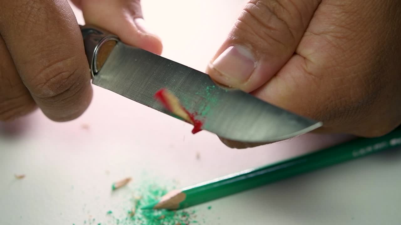 Footage of hands slowly sharpening a pencil and some coloured pencils with a sharp knife