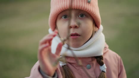 a front close-up of a little girl dressed in a pink jacket and beanie, intently examining and touching a dry tree branch. the image captures her curiosity and engagement with nature