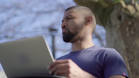 Bottom-view-of-young-man-in-park-opening-laptop,-thinking