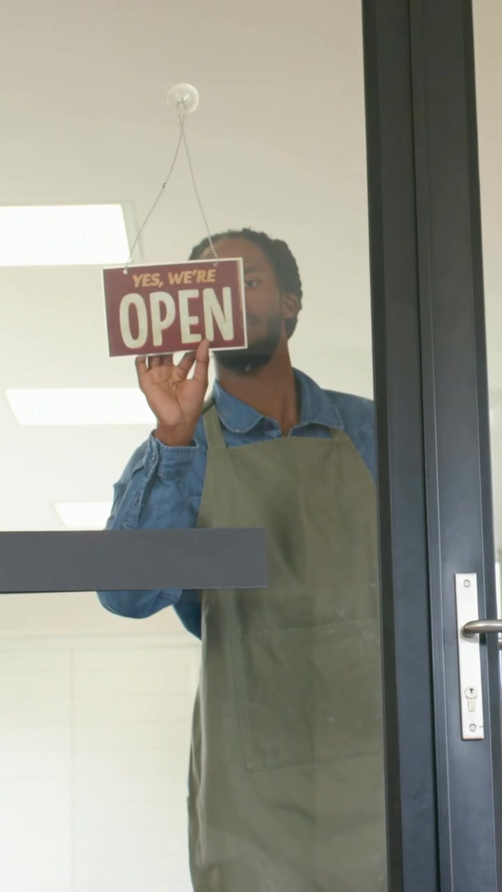Vertical video: Turning man in green apron changing sign to OPEN, pulling handle and opening door