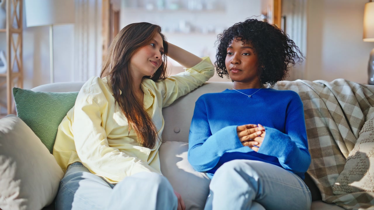 Two women sharing story living room couch. Diverse girlfriends bonding home