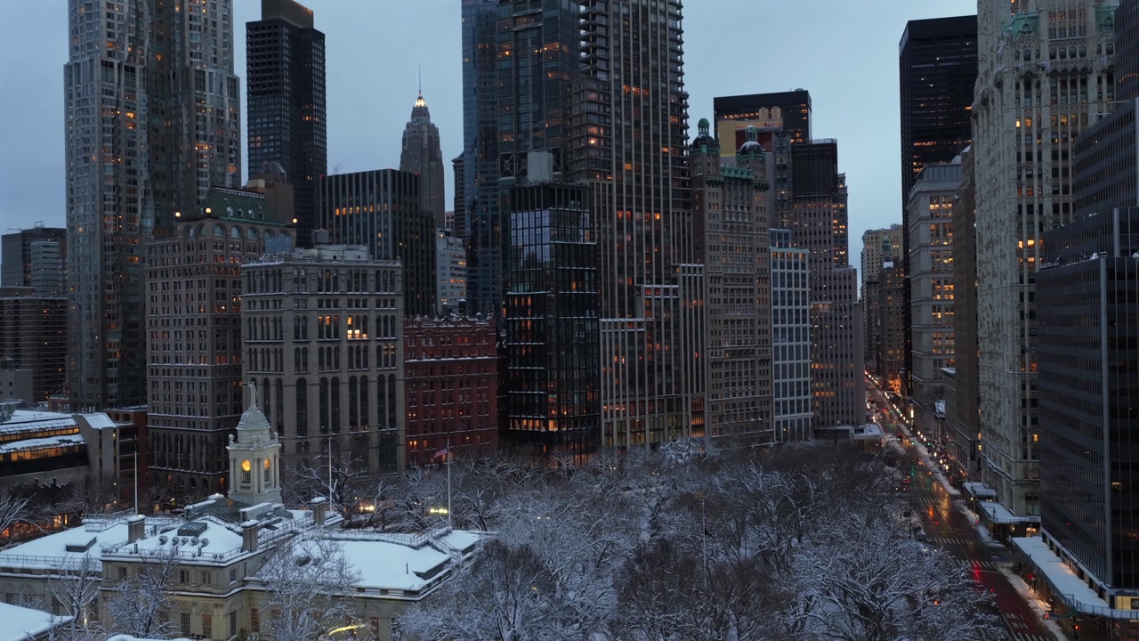 Aerial tilt up of New York City Hall and skyline at dusk in winter, snow covered park and streets below, glowing city lights, luxury towers and historic buildings in calm cold christmas atmosphere.
