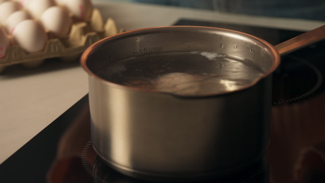 Hånd bruger ske til at lægge friskt æg i gryde af rustfrit stål med simrende vand - kogning af påskeæg. Nærbillede