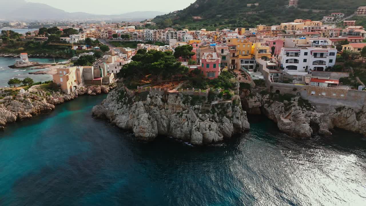 Drone pulls back and rises from a cliffside pink villa in Porticello, Sicily, revealing the coastal town, rocky shoreline, and vibrant architecture by the Tyrrhenian Sea