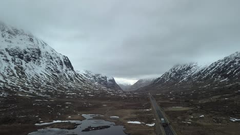 aerial backwards shot showing driving cars on road between scottish highlands during grey winter day - isle of skye, three sisters, scotland