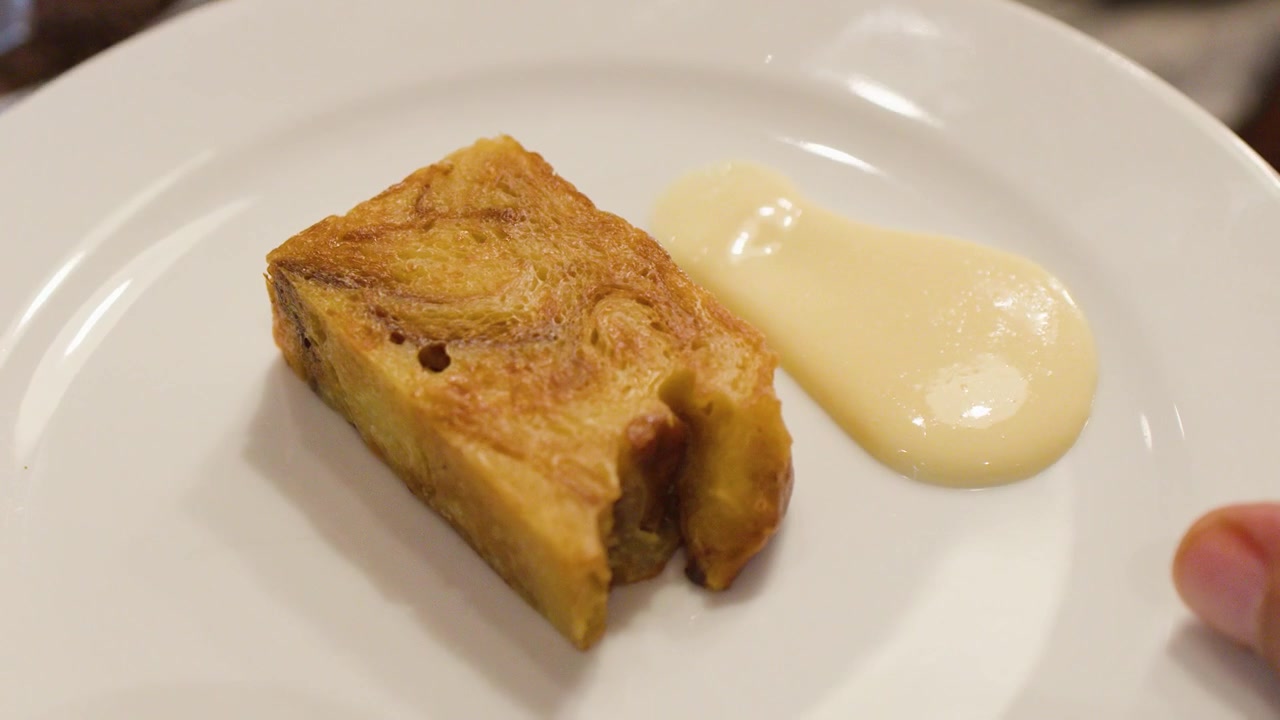 En metallskje heller glatt vaniljekrem ved siden av en skive varm brødpudding