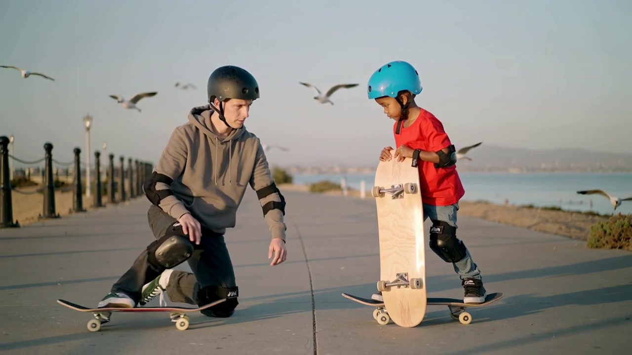 Starting with adult kneeling on skateboard, teaching child to balance on seaside promenade, helmets