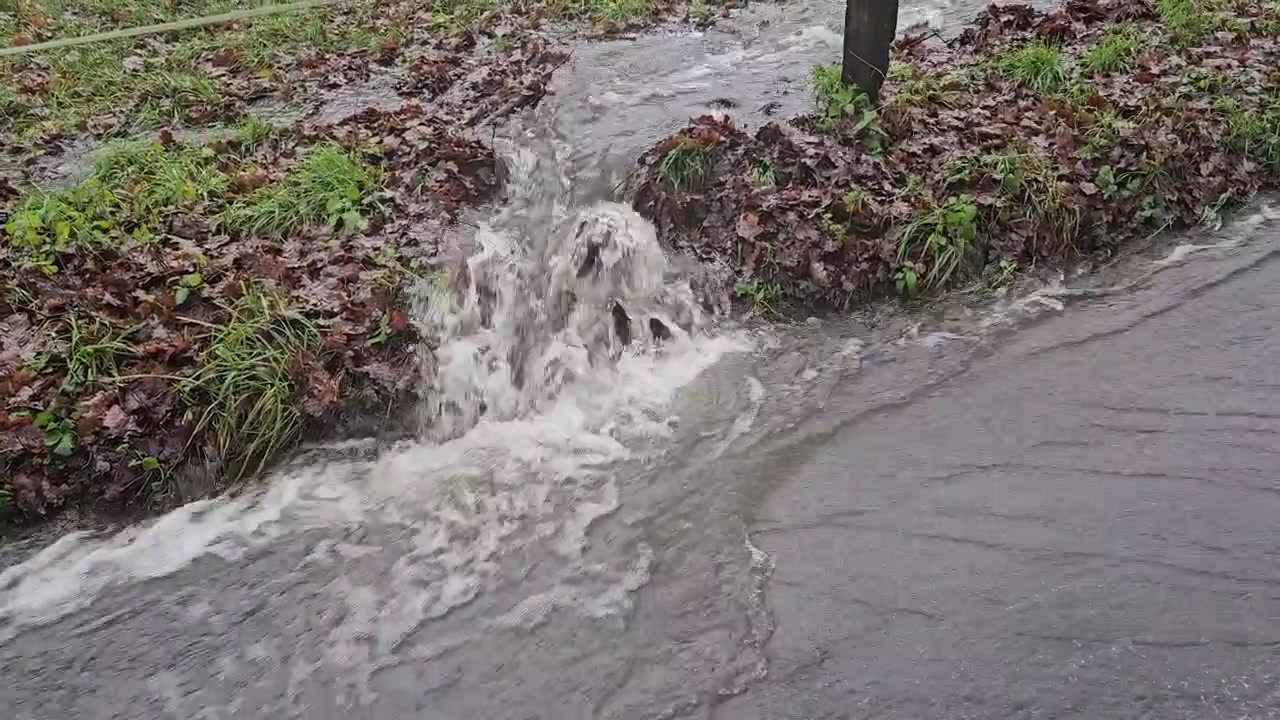 Floodwater from heavy rainfall gushing from a grassy bank and flowing rapidly onto a street