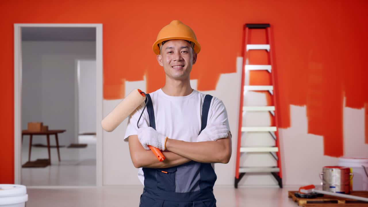 pintor hombre asiático con casco de seguridad sonriendo y cruzando los brazos mientras está de pie con la pared de pintura en la habitación