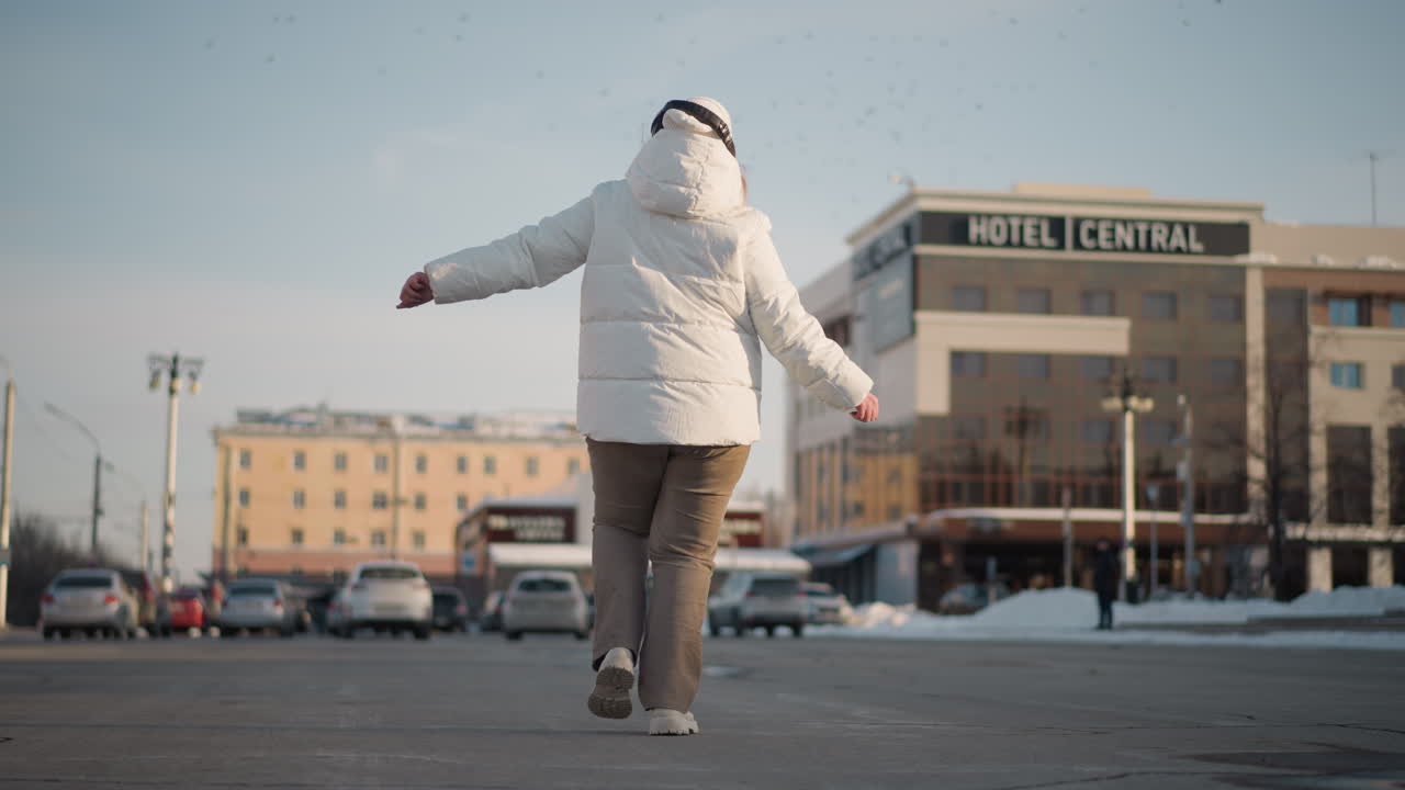 Back view of youth dancing confidently near car park wearing winter jacket and headphones, arms raised in expressive motion, surrounded by snowy pavement and hotel sign visible in background