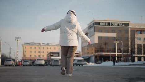 back view of youth dancing confidently near car park wearing winter jacket and headphones, arms raised in expressive motion, surrounded by snowy pavement and hotel sign visible in background