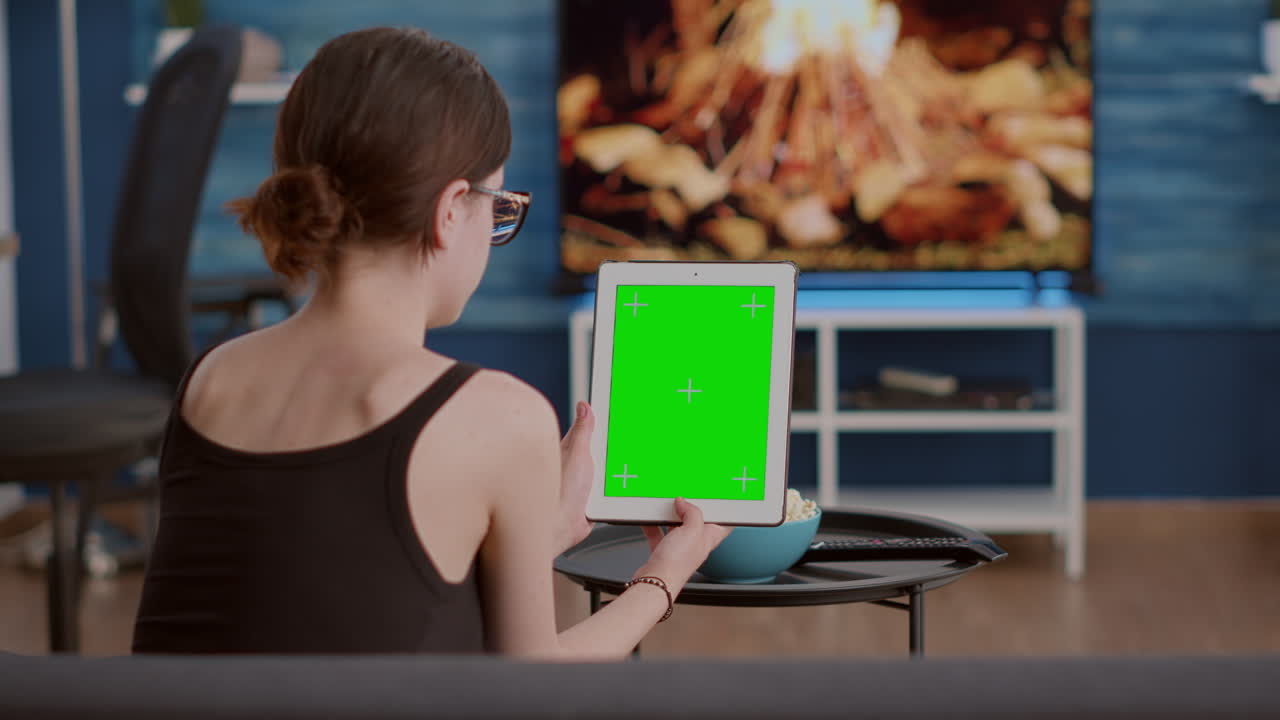 Young woman sitting on sofa holding vertical digital tablet with green screen watching online social media content