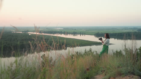 female photographer standing on hilltop capturing distant river landscape during golden hour with camera in hand, wild grass swaying gently, glass cup resting subtly in foreground among vegetation