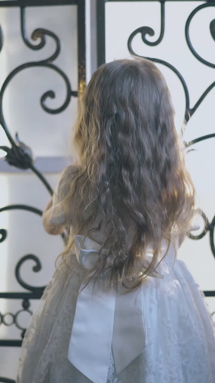 cute little blond girl in festive dress with bow opens beautiful forged lattice of room door at home backside view