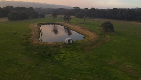 Aerial-orbit-shot-over-mystic-house-and-lake-during-a-foggy-sunset-at-farm