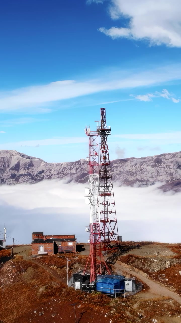 Trasmettitore in vetta alla montagna sopra le nuvole, drone aereo rivela una torre di telecomunicazioni color ruggine su un crinale brullo.