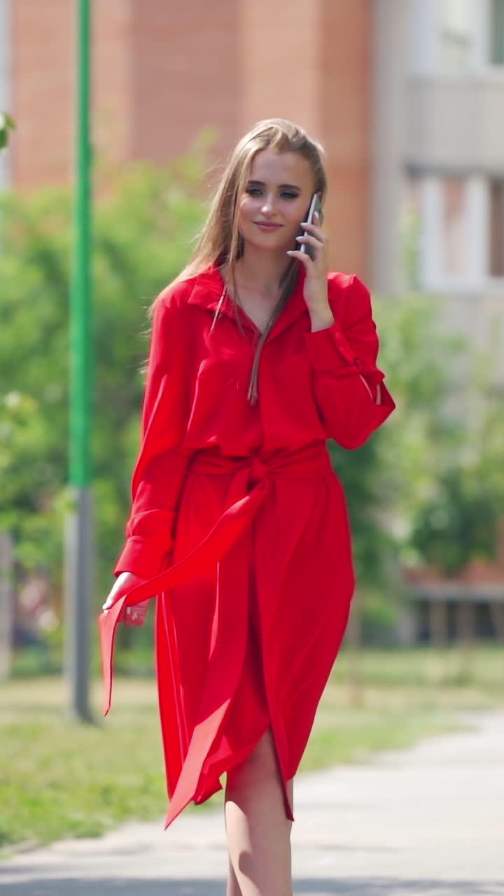 Sexy woman in red dress outdoors. Young lady walking slowly on the street and talking on the mobile phone. Beautiful female in passionate dress in a summertime. Vertical video
