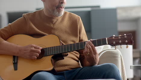 focused senior biracial man sitting on couch playing guitar at home, slow motion