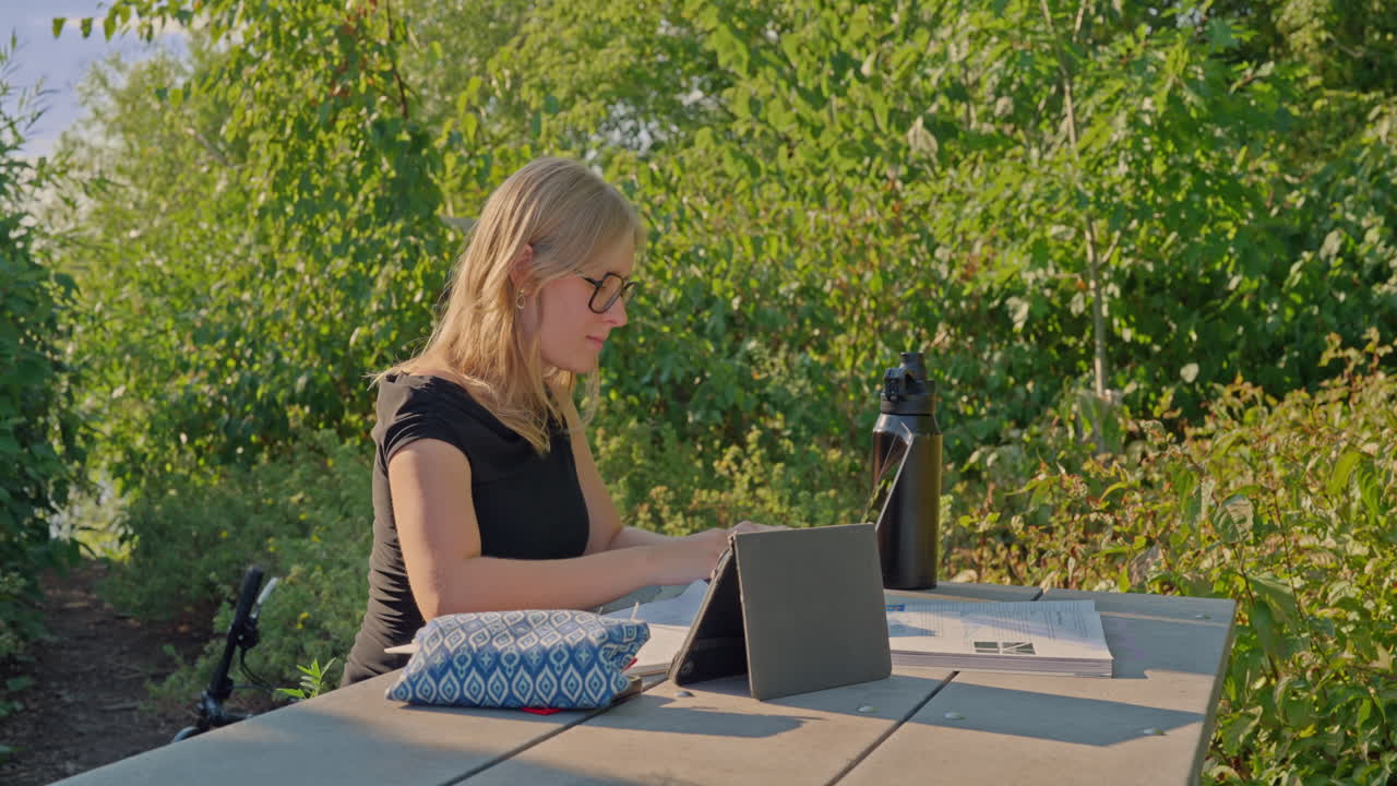 En studerende fra Generation Z med briller, der studerer alene udendørs med en tablet, notesbog og genanvendelig vandflaske på et bord i parken. Dolly-kamerakørsel mod venstre, konceptet digital læring og bæredygtighed