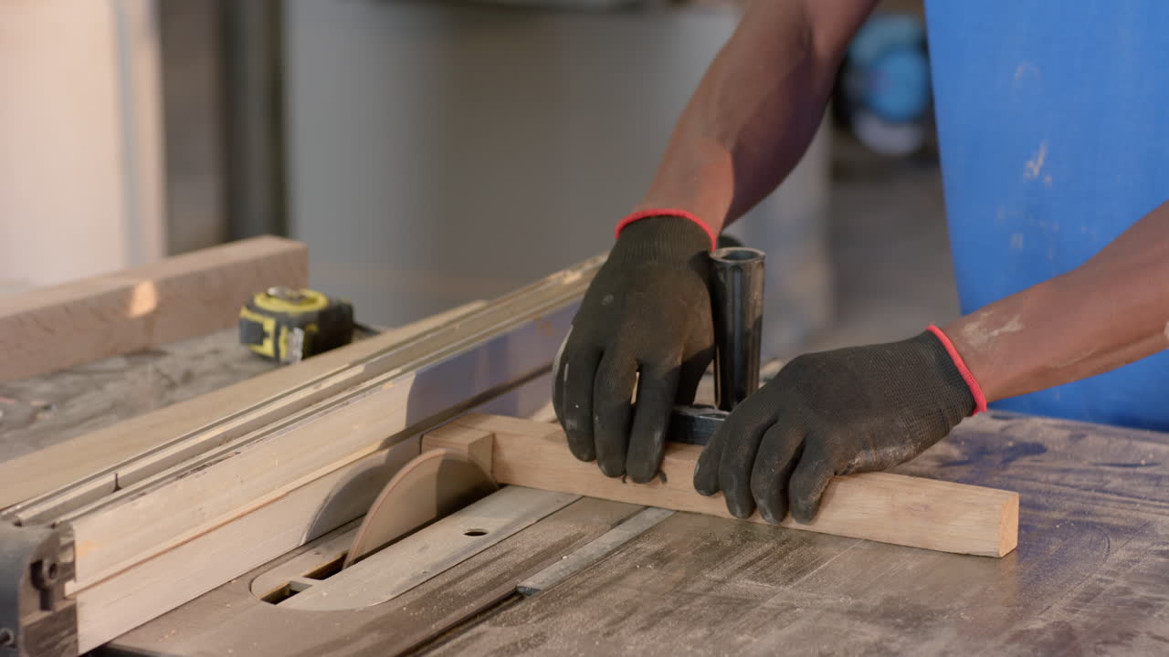 African American man guiding wood plank through table saw in shop, wearing gloves, copy space