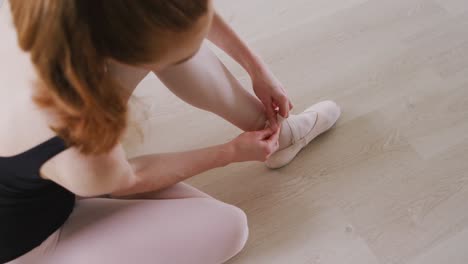 caucasian female ballet dancer sitting on the floor in the studio and tying her ballet shoes