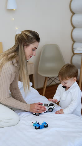 long-haired woman sits on the bed and little baby stands beside. mom fixes a toy car for her son. vertical video.