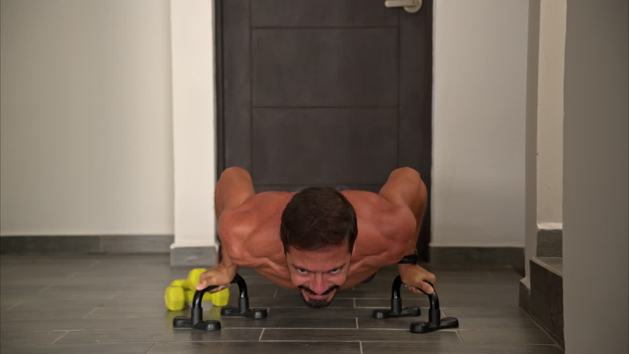 vista frontal en cámara lenta de un hombre latino hispano en forma entrenando en casa haciendo flexiones usando barras de apoyo de ayuda
