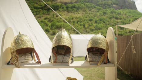 A slow pull-out shot reveals a display of ancient warrior equipment at a reenactment camp. The shelves hold ornate bronze helmets, armor, and other historical artifacts
