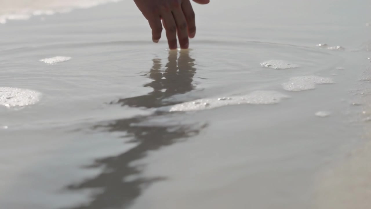 Lowering person's hand touching seawater at shoreline, making ripples and distorting reflection