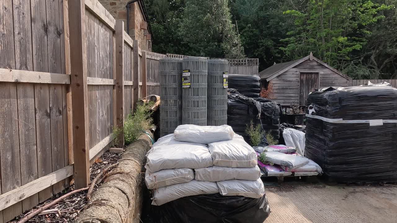 A white utility van enters an outdoor supply yard with stacked fertilizer bags, machinery, and storage bins under daylight, surrounded by greenery and wooden fencing