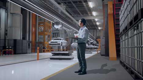 vista lateral completa del cuerpo de un trabajador profesional masculino asiático de pie en una fábrica de automóviles ev, instalación industrial de alta tecnología, observa mirando hacia arriba antes de llegar a concentrarse en la computadora portátil y seguir escribiendo