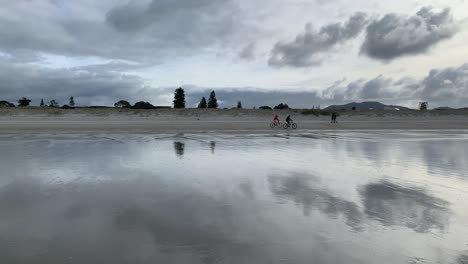 A-pair-of-cyclists-rode-passed-two-walkers-and-a-dog-on-the-beach-under-a-cloudy-sky-in-Omaha-Warkworth-New-Zealand