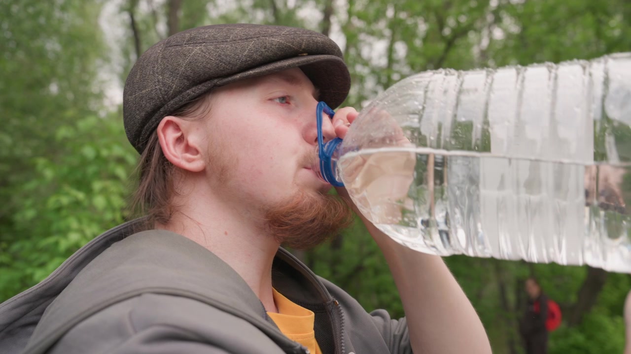 Primo piano di un uomo che beve acqua da una grande bottiglia di plastica in un parco. L'uomo porta alla bocca un contenitore trasparente coperto di condensa, indossando un berretto piatto e una giacca, con lo sfondo visibile.