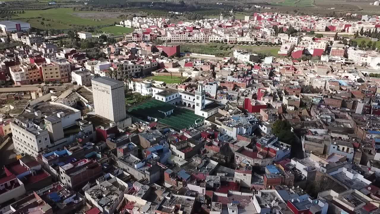 Side-angle drone shot capturing the intricate architecture of Larache's Grand Mosque. Highlights the historic minaret, inner courtyard, and the surrounding traditional Moroccan city dwellings
