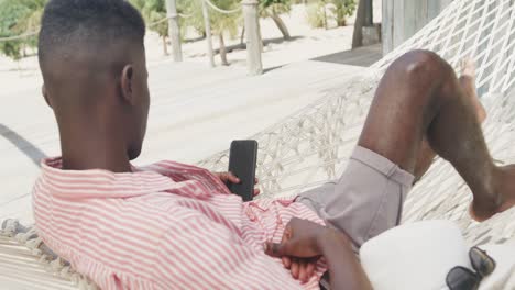 hombre afroamericano acostado en una hamaca usando un teléfono inteligente en una playa soleada, cámara lenta