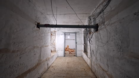 movement by a long corridor with bare walls and some wires hanging from the ceiling. entering the well-lit room with added brick wall in the middle.