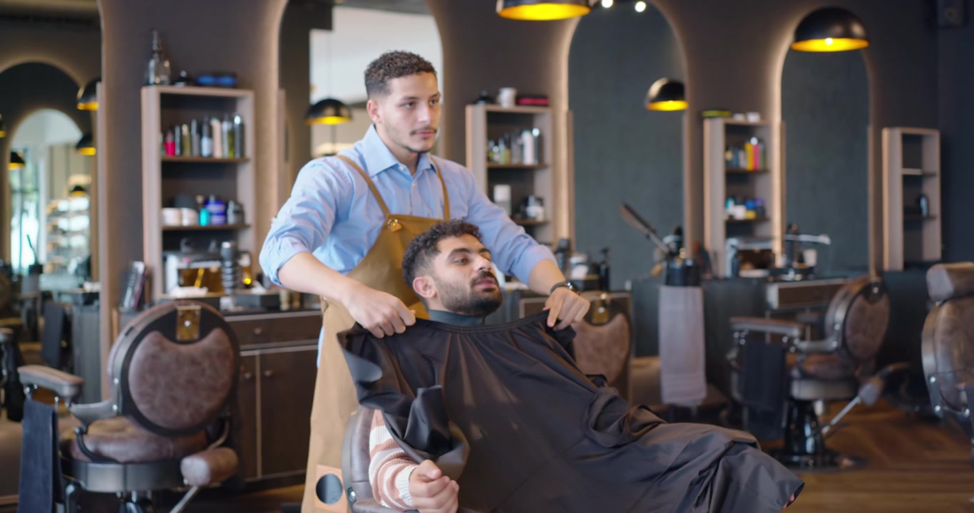 En mandlig frisør sætter halsstrimmel på og lægger kappe over en kunde i en barbershop, mens den mandlige kunde venter på en klipning.