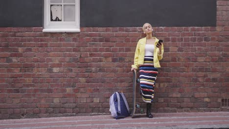 Mixed-race-woman-leaning-against-a-brick-wall-with-her-skateboard-and-her-phone