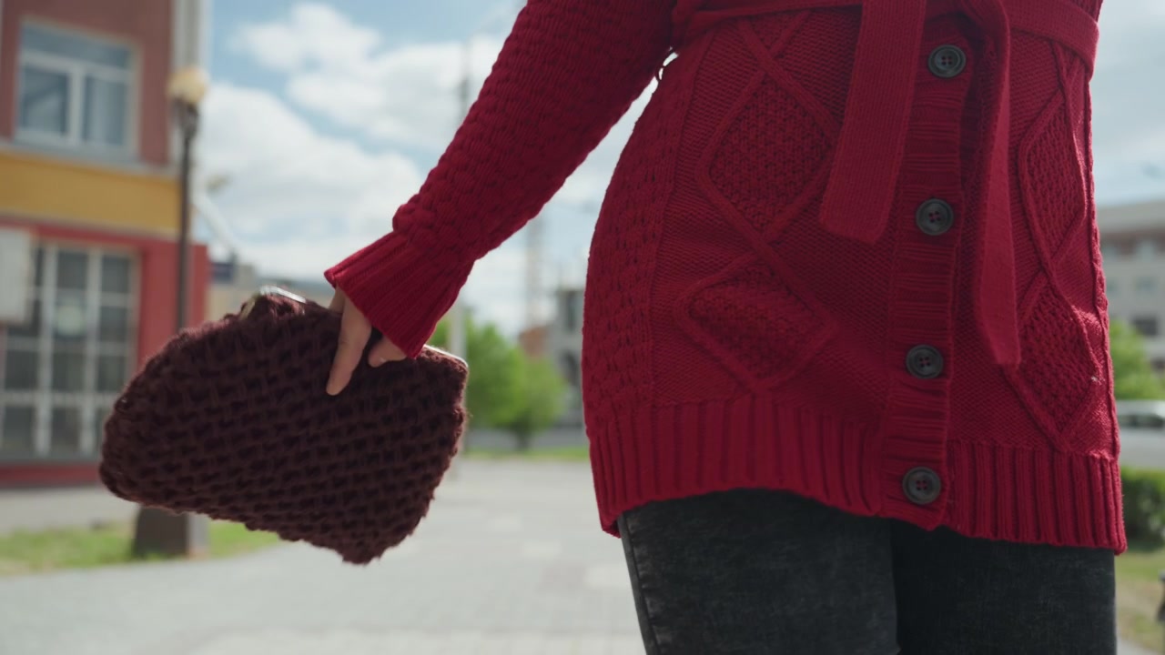 Close-up de mulher com cardigã de tricô vermelho caminhando ao ar livre, segurando uma bolsa de mão de tricô marrom. A cena mostra um fundo urbano com um prédio, poste de luz e iluminação suave da luz do dia. A câmera permanece